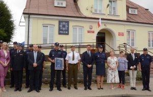 Umundurowani policjanci oraz osoby cywilne stojące przed budynkiem posterunku