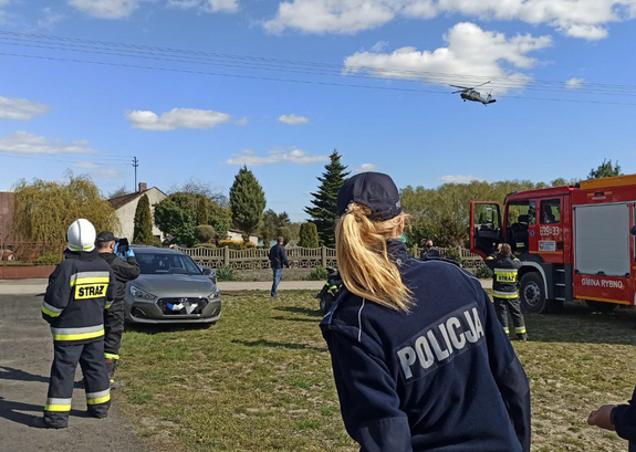 Policjanci i strażacy stojący na łące, w tle policyjny śmigłowiec