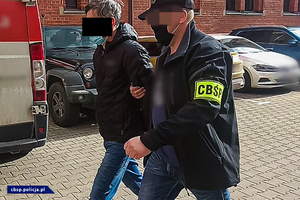 Policjant w kurtce z napisem &quot;CBŚP&quot; prowadzący zatrzymanego.