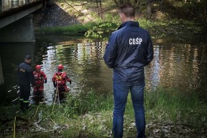 Policjant w kurtce z napisem &amp;quot;CBŚP&amp;quot;, w tle strażacy wchodzący do rzeki.