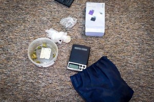 Materiałowy woreczek, waga elektroniczna oraz plastikowe, okrągłe pudełko wypełnione suszem roślinnym leżące na wykładzinie podłogowej.