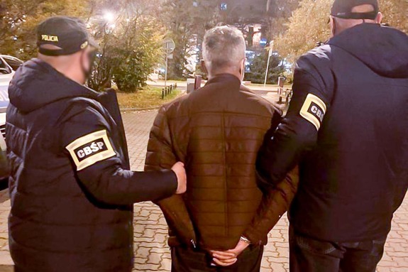 Policjanci w strojach cywilnych. Na ramionach mają opaski z napisem CBŚP. W środku pomiędzy nimi zatrzymany mężczyzna w kajdankach założonych na ręce trzymane s tyłu.