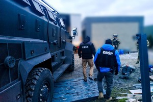 Policjanci z oznaczeniami &quot;CBŚP&quot; wchodzą na teren posesji na której leżą zatrzymane osoby nad którymi stoją policyjni kontrterroryści. Z lewej strony policyjny transporter.