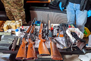 Różnego rodzaju broń myśliwska, pistolet maszynowy, broń krótka, magazynki, tłumiki, amunicja w opakowaniach leżące na blacie stołu.