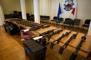 Różnego rodzaju broń długa leżąca na blacie stołu. W tle na ścianie logotyp CBŚP, flaga Polski i UE.