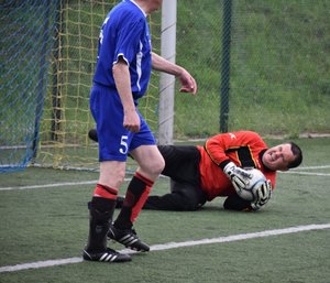 Fotografia kolorowa na której widać lezącego na boisku bramkarza, który w rękach ma piłkę. Przed nim stoi zawodnik ubrany w niebieski strój sportowy.