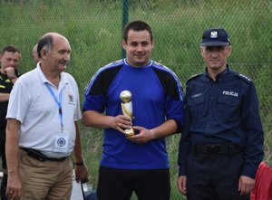 Fotografia kolorowa na której w środku znajduje się zawodnik- bramkarz z pucharem w rekach. Po jego lewej stronie stoi przedstawiciel IPA a po prawej Komendant Komisariatu Policji w Pruchniku.