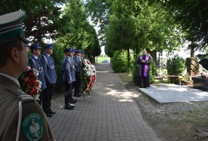 Fotografia kolorowa przedstawiająca alejkę na cmentarzu. Na zdjęciu widać umundurowanych policjantów oraz funkcjonariusza Straży Granicznej. Po prawej stronie zdjęcia, przy nagrobku stoi ksiądz.