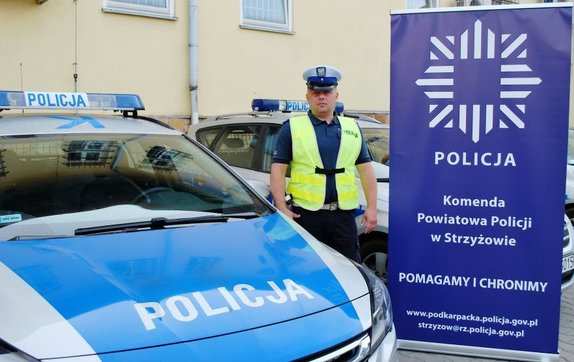 Policjant strzyżowskiej drogówki który pomógł choremu. W tle radiowóz oraz baner strzyżowskiej policji z hasłem &quot;Pomagamy i chronimy&quot;