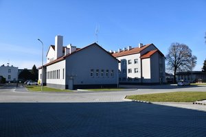 Widok z tyłu na prawe i lewe skrzydło komendy oraz fragment jej części środkowej po remoncie. Na pierwszym planie trawnik i plac wyłożony szarą kostką brukową. Po lewej stronie brama wjazdowa i parking przy komendzie.