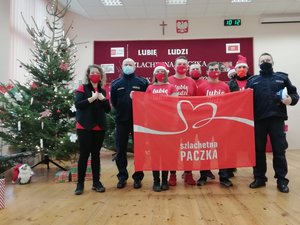 Kolorowa fotografia. W magazynie szlachetnej paczki, na scenie wspólnie stoją wolontariusze i policjanci, wśród nich komendant powiatowy. Z boku choinka. Wolontariusze i policjanci trzymają baner z logo szlachetnej paczki.