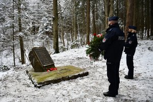 Złożenie kwiatów i zapalenie zniczy przy obelisku upamiętniającym katastrofę śmigłowca w Cisnej przez policjantów. Policjantka i policjant stoją przed obeliskiem z wieńcem i zniczami.