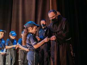 Kolorowa fotografia. Ojciec kustosz na scenie wręcza jednemu z małych policjantów legitymację słuchacza.