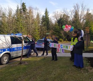 Na zdjęciu widać trzy policjantki, policyjnego misia, siostrę zakonną i dwa radiowozy z włączonymi sygnałami błyskowymi. Jedna z policjantek z siostrą zakonną trzymają transparent z napisem &quot;Uśmiechnij się&quot;.