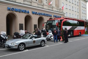 Przedstawiciele służb mundurowych oczekują na rejestrację przed krwiobusem. Na pierwszym planie zaparkowane przed komendą motocykle i kabriolet, w tle po prawej krwiobus.