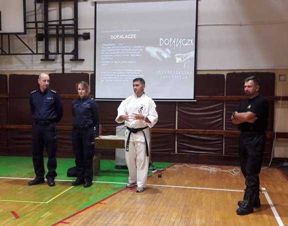 Na fotografii asp. Grzegorz Uzar w kimono karate oraz kilku innych policjantów, w tle widać również rzutnik na którym wyświetlana jest prezentacja multimedialna dotycząca tematu dopalaczy. Spotkanie zorganizowane przez mł. asp. Grzegorza Uzara „Przestępczość narkotykowa dzieci i młodzieży – stop uzależnieniom”