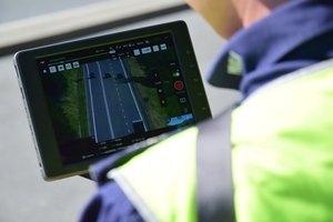Zbliżenie na panel sterowania dronem. Na ekranie widoczna droga (z góry, z kamery drona). Policjant trzyma panel w rękach.