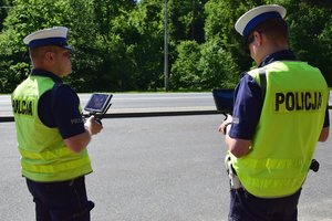 Dwaj policjanci ruchu drogowego, umundurowani i w odblaskowych kamizelkach, trzymają w rękach panel sterowania dronem.