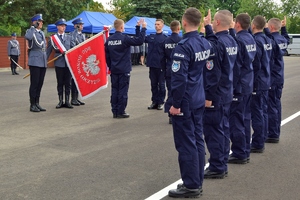 Sztandar po lewej stronie, po prawej nowo przyjęci funkcjonariusze podczas ślubowania. Mają uniesione prawe dłonie i dwa palce w geście ślubowania.