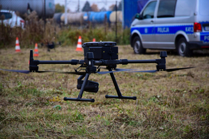 Policyjny dron nad ziemią. W tle policyjny radiowóz