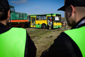 Na środku w tle autobus - strażacy w środku i na zewnątrz udzielają pomocy poszkodowanym. Po prawej i lewej w pierwszym planie widoczne sylwetki strażaków w kamizelkach odblaskowych