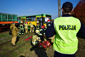 Na środku w tle autobus - strażacy w środku i na zewnątrz udzielają pomocy poszkodowanym. Na pierwszym planie policjant w kamizelce odblaskowej