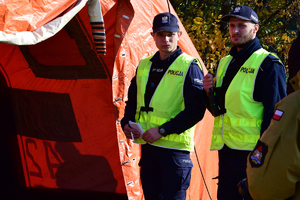 Służby medyczne w pomarańczowym namiocie udzielają pomocy poszkodowanym. Na pierwszym planie po prawej policjanci w kamizelkach odblaskowych