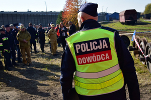 Na pierwszym planie policjant w furażerce i kamizelce odblaskowej z napisem POLICJA DOWÓDCA. W tle policjanci i przedstawiciele służb i instytucji