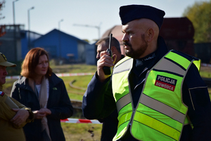 Na pierwszym planie policjant w furażerce i kamizelce odblaskowej z napisem POLICJA DOWÓDCA, rozmawia przez krótkofalówkę. W tle Wojewoda Podkarpacki Ewa Leniart