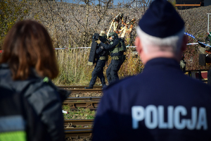 Policyjni antyterroryści idą w szeregu wzdłuż torowiska. Pierwszy z lewej trzyma tarczę ochronną. Na pierwszym planie dwie niewyraźne sylwetki odwrócone plecami