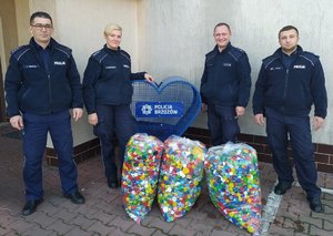 Zdjęcie przedstawia czterech umundurowanych policjantów (w tym jedna policjantka) stojących przy opróżnionym niebieskim sercu na nakrętki. Przed nimi stoją trzy pełen worki nakrętek.