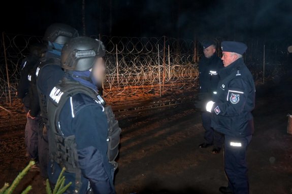 Po prawej na pierwszym planie Komendant Wojewódzki Policji w Rzeszowie, po lewej policjanci pełniący służbę na granicy. W tle widoczne zabezpieczenia graniczne. Zdjęcie wykonane w nocy.