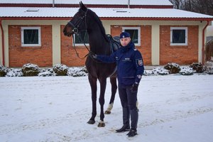 Policjant stoi obok konia, trzyma go za wodze. Zdjęcie wykonane od przodu w ciągu dnia, w tle stajnie, padający śnieg.