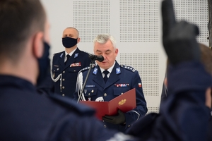 Na środku Komendant Wojewódzki Policji w Rzeszowie przy mikrofonie. W rękach trzyma ozdobną oprawę ze złotym godłem Polski. W tle kadra kierownicza podkarpackiej Policji.