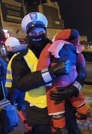 Zdjęcie kolorowe wykonane w porze nocnej-przedstawia policjanta umundurowanego , który trzyma małe dziecko na rękach dziecko ubrane jest w kombinezon w kolorze czerwonym i z dodatkami niebieskimi