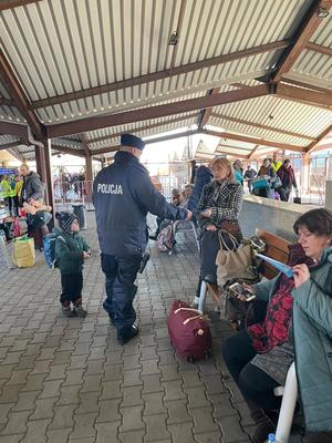 Zdjęcie kolorowe wykonane w porze dziennej  – Przedstawia stację PKP w Przemyślu a w tle widoczny policjant rozdający uchodźcom ulotki