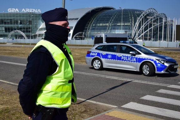 Na pierwszym planie policjant oddziału prewencji w furażerce. W tle policyjny radiowóz oraz budynek G2A Arena.