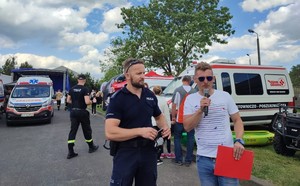 policjant udziela wywiadu podczas pikniku charytatywnego. W tle pojazdy ratownicze