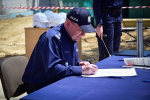 Policjant przy stole podpisuje akt erekcyjny. W tle plac budowy