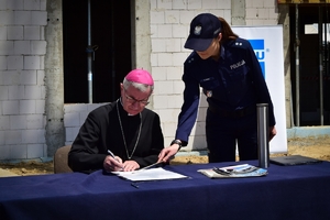 Biskup diecezji rzeszowskiej przy stole podpisuje akt erekcyjny. W tle plac budowy