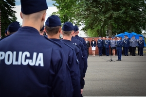 Po lewej na pierwszym planie nowo przyjęci policjanci (widok od tyłu, na mundurach napis POLICJA), w tle Komendant Wojewódzki Policji w Rzeszowie przy mikrofonie, dalej w tle zaproszeni goście i kadra kierownicza KWP.