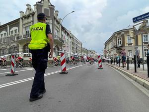 Po lewej policjant zabezpieczający przejazd kolarzy. W tle kolarze przejeżdżający ulicami.