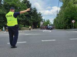 Na pierwszym planie (po lewej) policjant stojący na ulicy i wskazujący kierunek kolarzom. W tle przejeżdżający peleton kolarski.