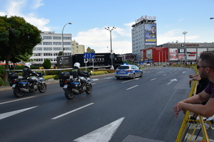 Policyjny radiowóz jadący ulicą, za nim dwa policyjne motocykle. Widok od tyłu.