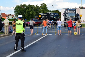 Po lewej stronie policjant stojący przy ulicy (widok od tyłu). W tle kibice i przejeżdżający ulicą kolarze.