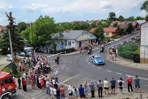 Policyjny radiowóz na zakręcie, za nim motocykle. Wokół drogi kibice i stojący wóz strażacki.