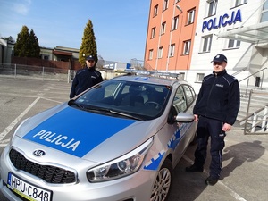 Na zdjęciu dwaj policjanci w granatowych mundurach stojący po obu stronach oznakowanego radiowozu