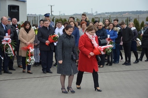 Dwie kobiety, z których jedna trzyma kwiaty. Za nimi pozostali uczestnicy uroczystości w cywilnych strojach.