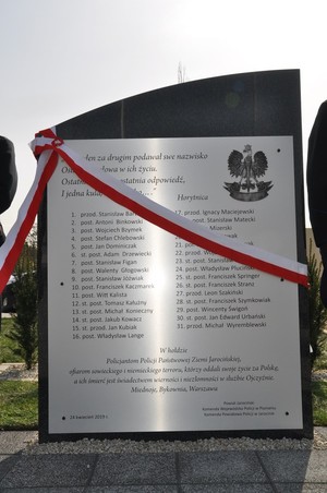 Obelisk z pamiątkową tablicą z nazwiskami pomordowanych policjantów z biało czerwoną szarfą