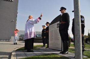 Ksiądz z kropidłem święci obelisk obok, którego stoją policjanci w mundurach z epoki międzywojennej. W tle kobieta robi zdjęcie.